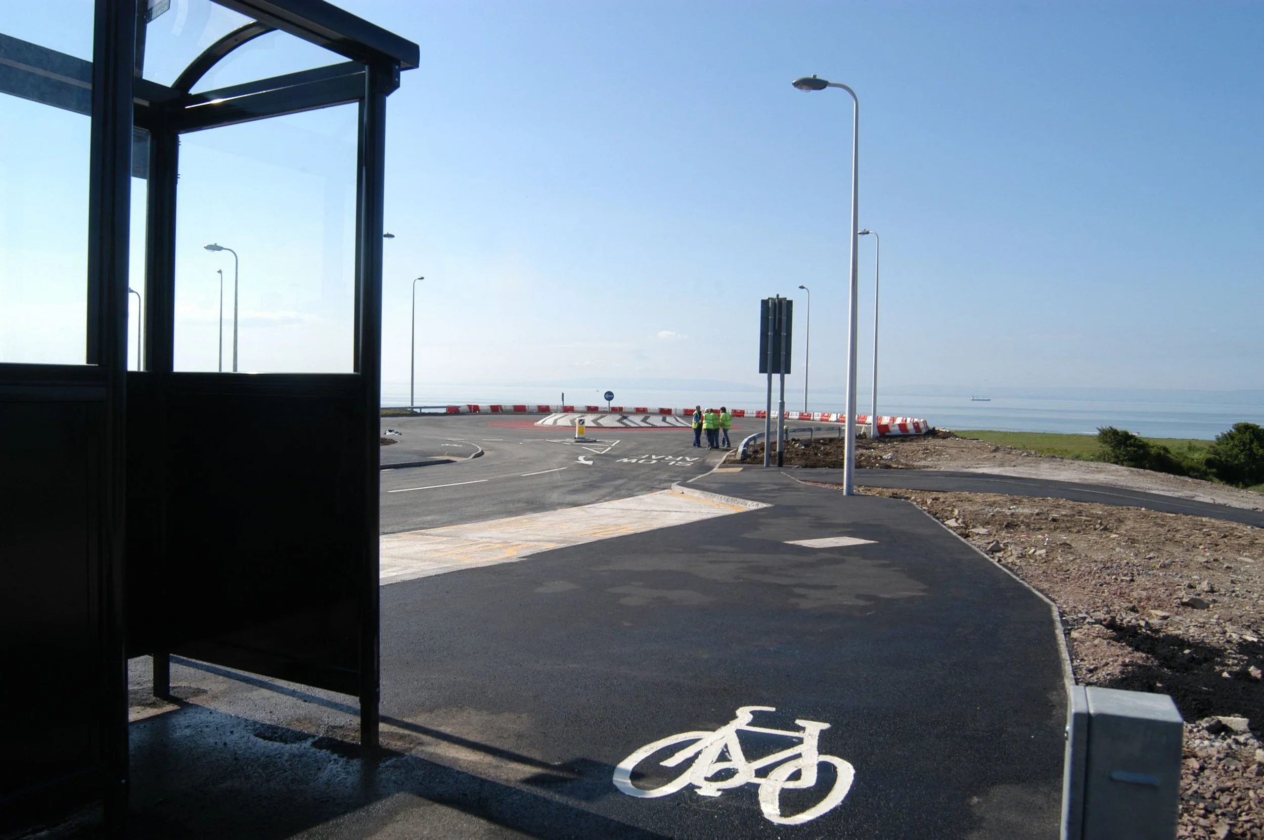 A bike lane painted on a new paved road leading towards a roundabout, with a bicycle symbol visible in the foreground, and a bus stop shelter on the left side of the image.