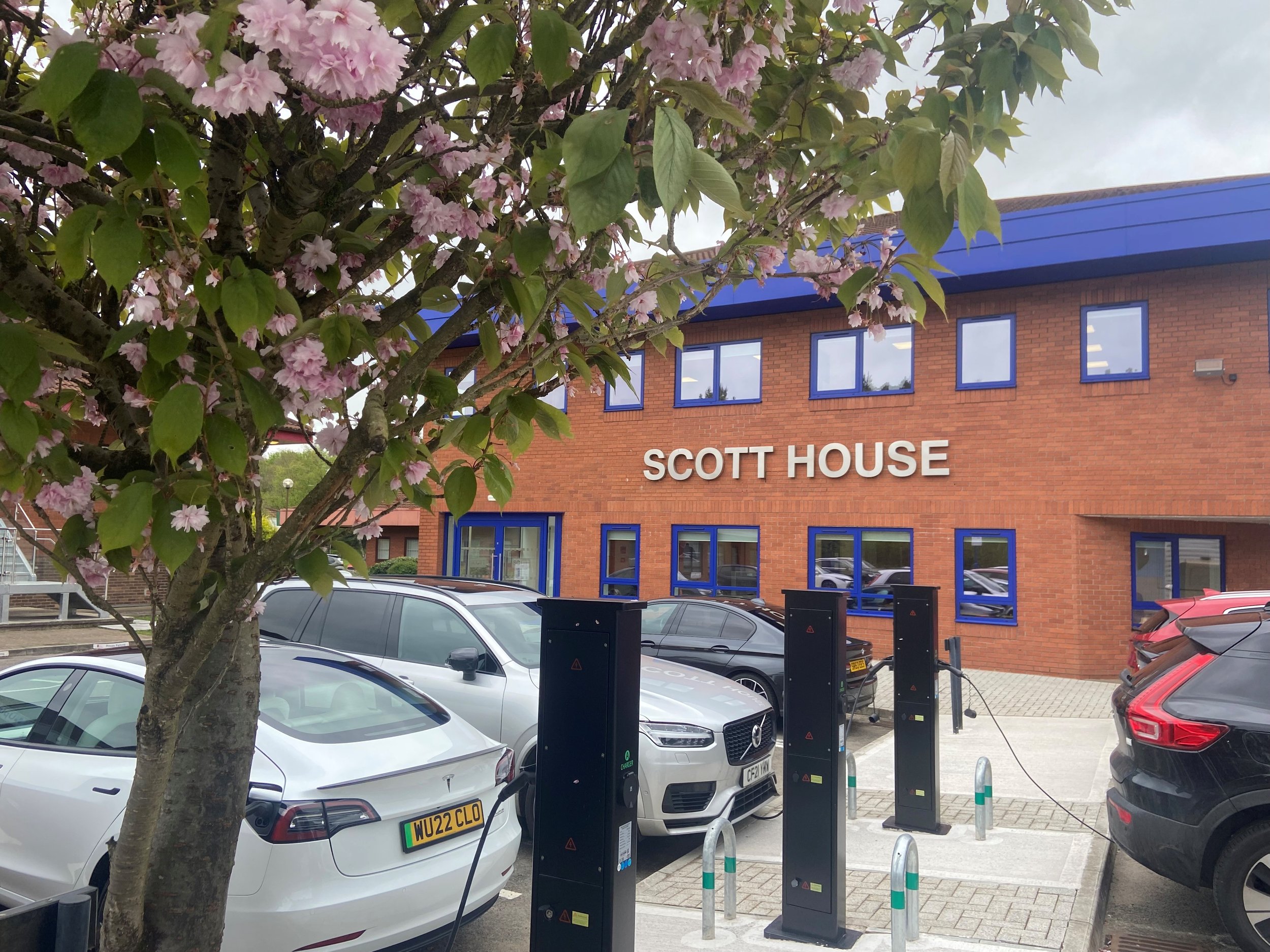 Building with a sign that reads 'Scott House', parked cars, electric vehicle charging stations, and a blossoming pink flowered tree in the foreground.