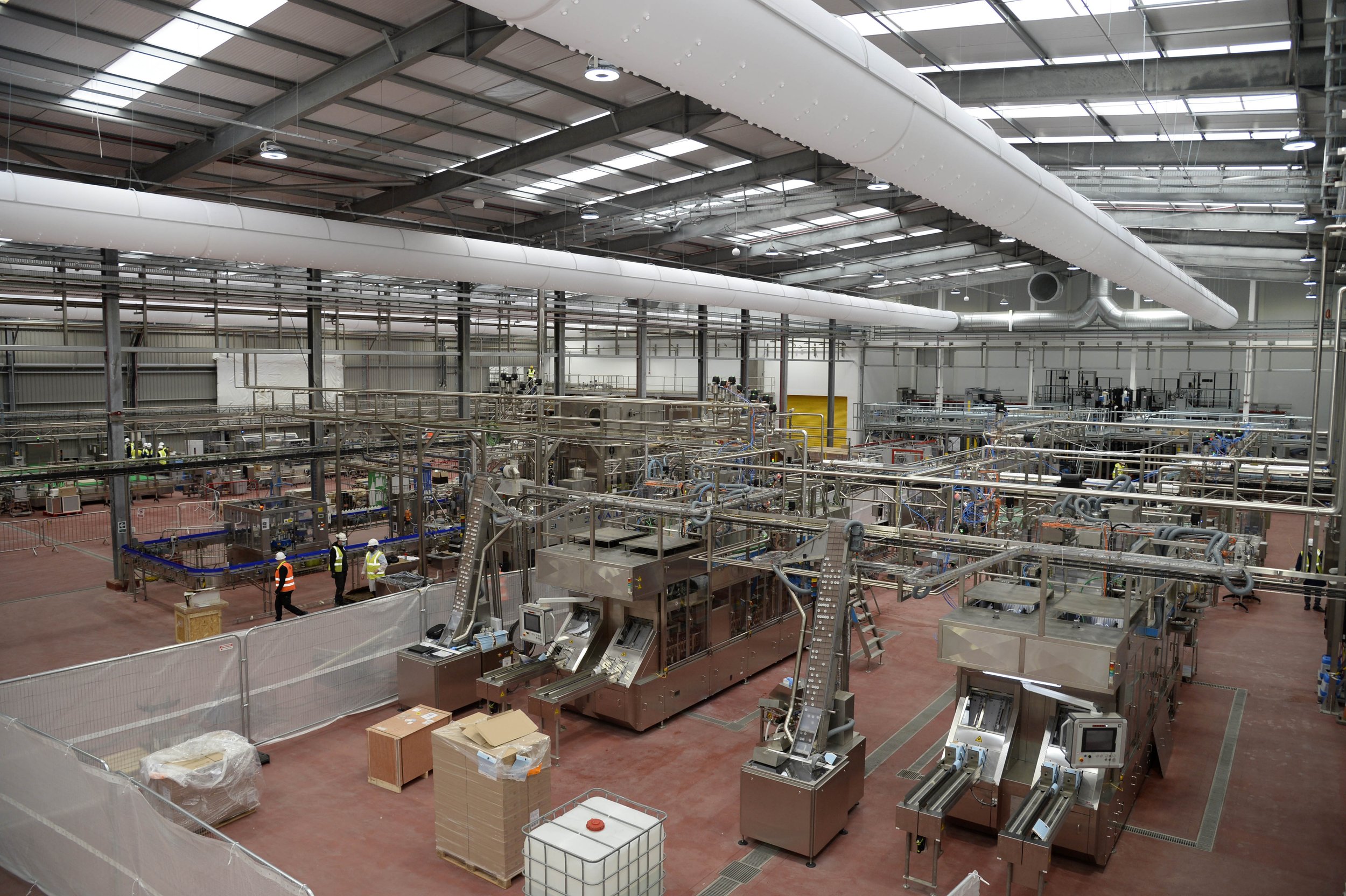 Industrial factory interior with machinery, conveyor belts, and workers in safety vests.