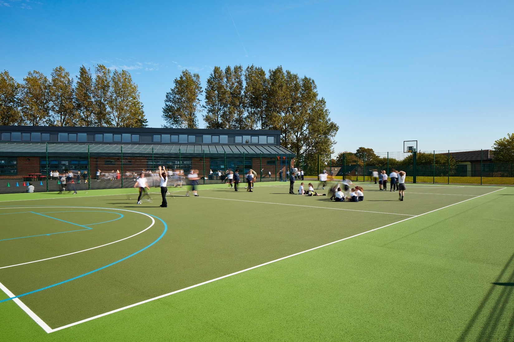 Children playing on an outdoor school sports court with a building and trees in the background under a clear blue sky.
