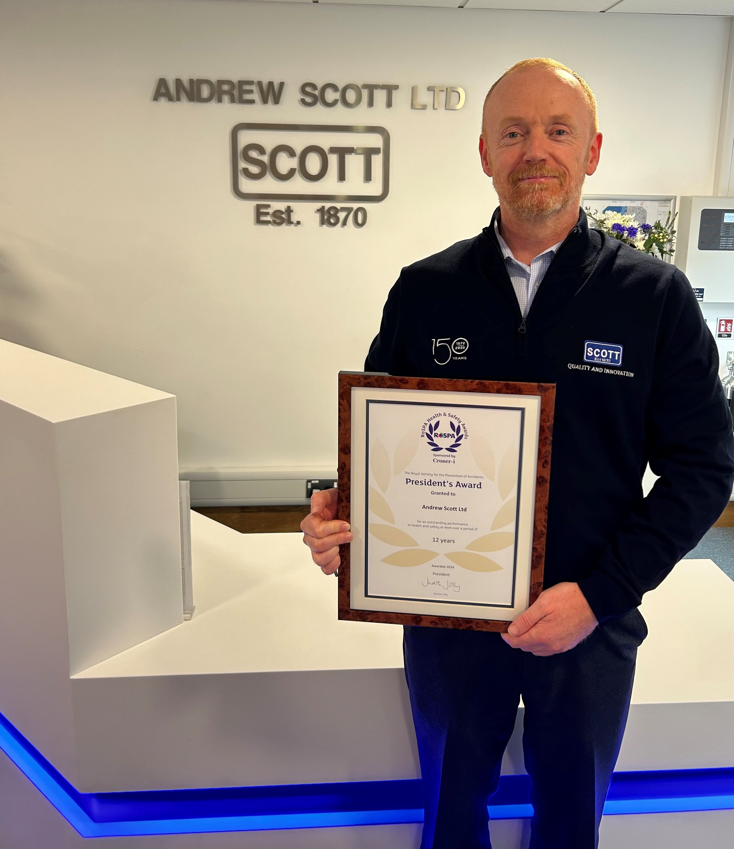 A man holding a framed award certificate in an office lobby. The background shows a wall with the logo and name "Andrew Scott LTD" and a reception desk with blue lighting.