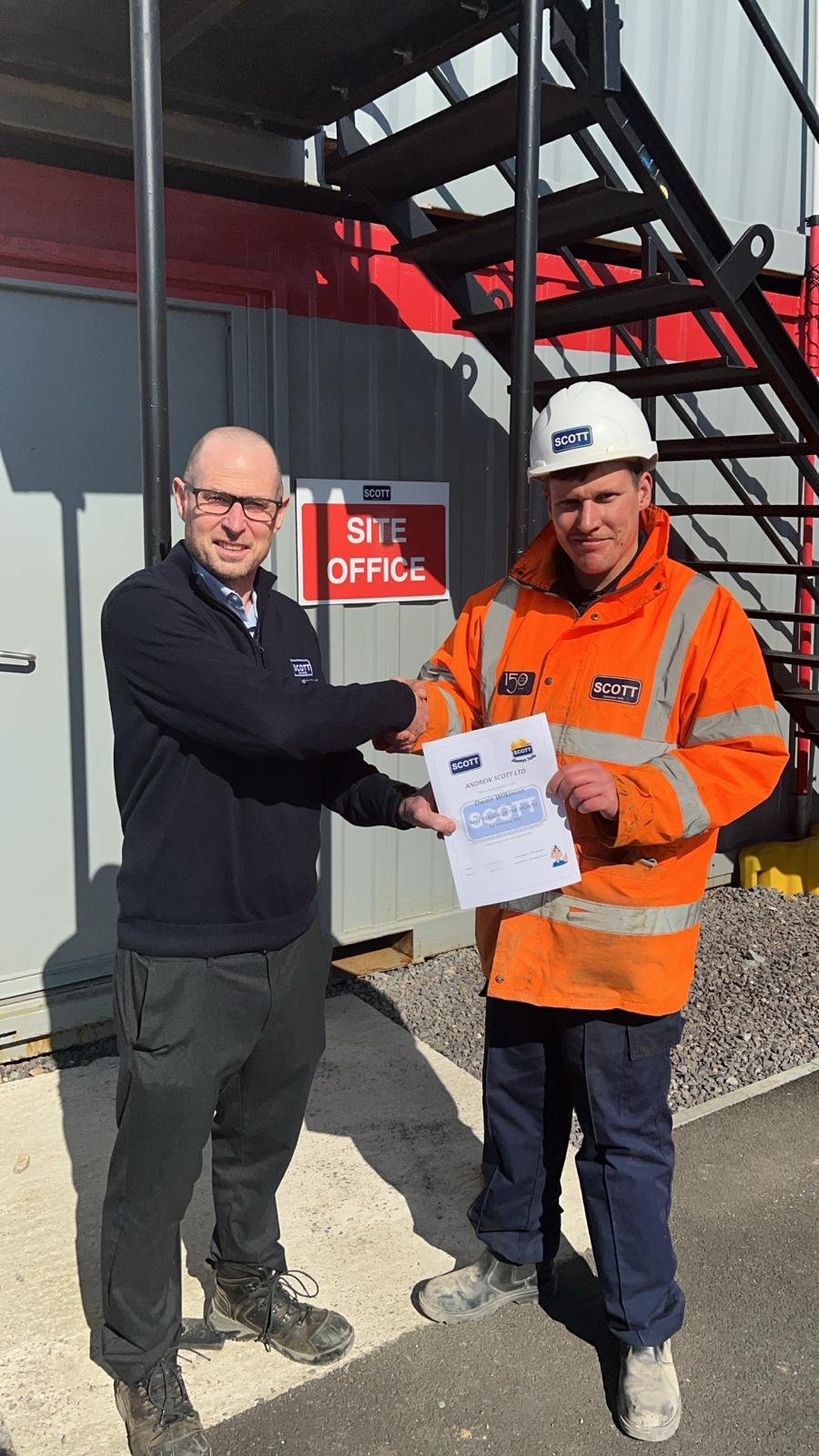Two men shaking hands outside a construction site. One man is wearing a black jacket and glasses, while the other is dressed in an orange safety jacket and a white hard hat. The man in safety gear is holding a certificate. Behind them is a building w