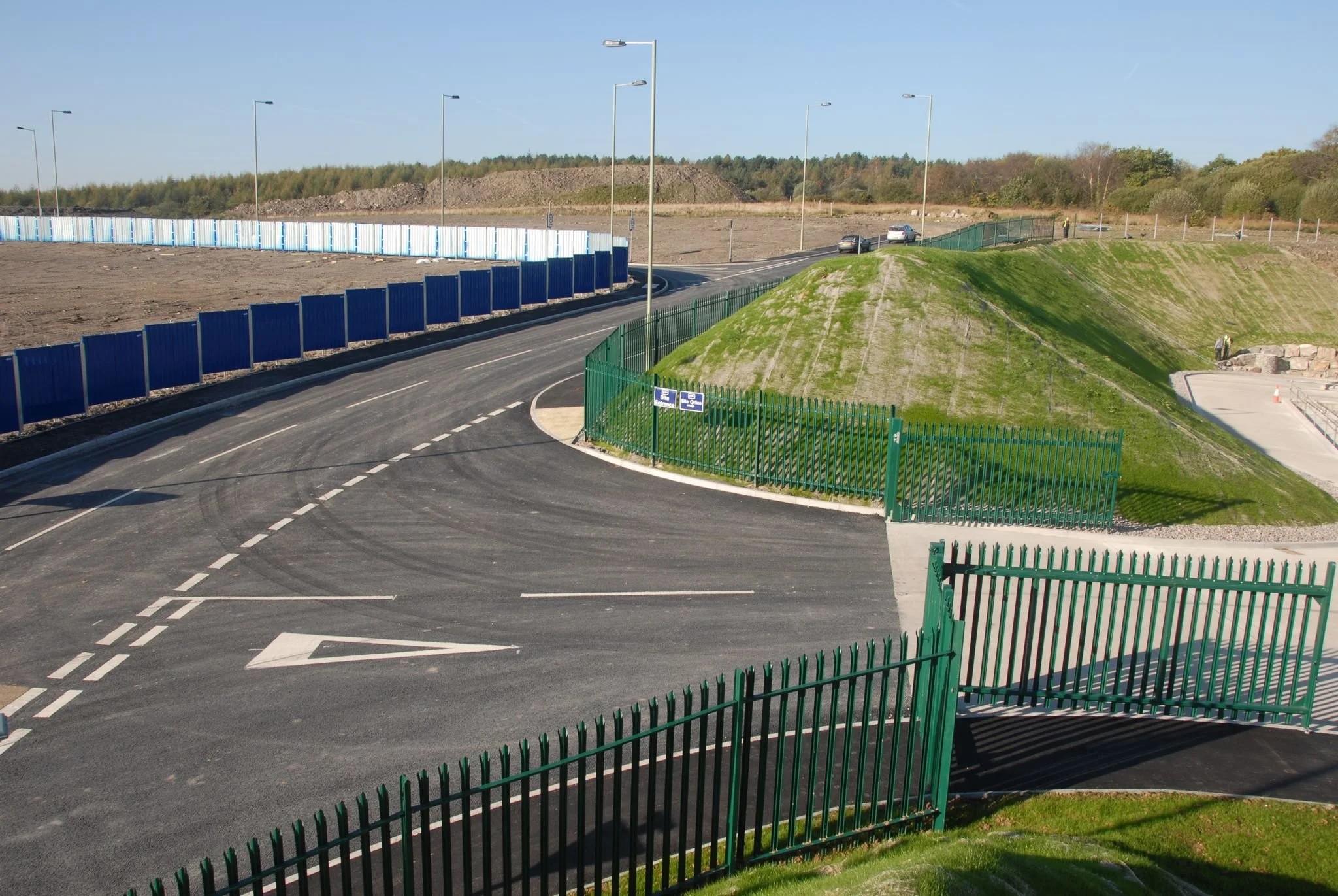 Empty paved parking lot with green metal fence, a grassy mound, and a blue sound barrier fence in the background under a clear sky.