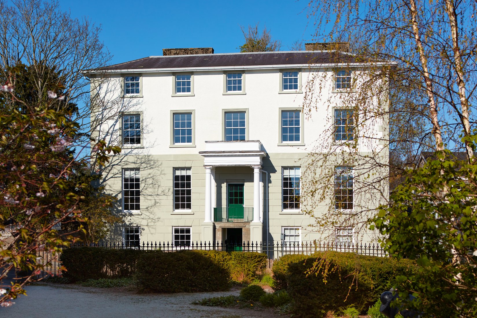 Georgian-style building with white facade, green door, and large windows, surrounded by trees and bushes, set against a clear blue sky.