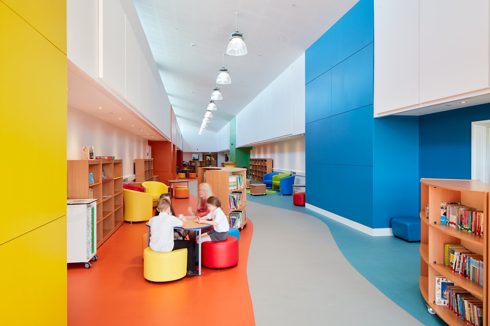 Brightly colored library interior with children sitting at a round table, bookshelves with colorful books, and modern furniture.