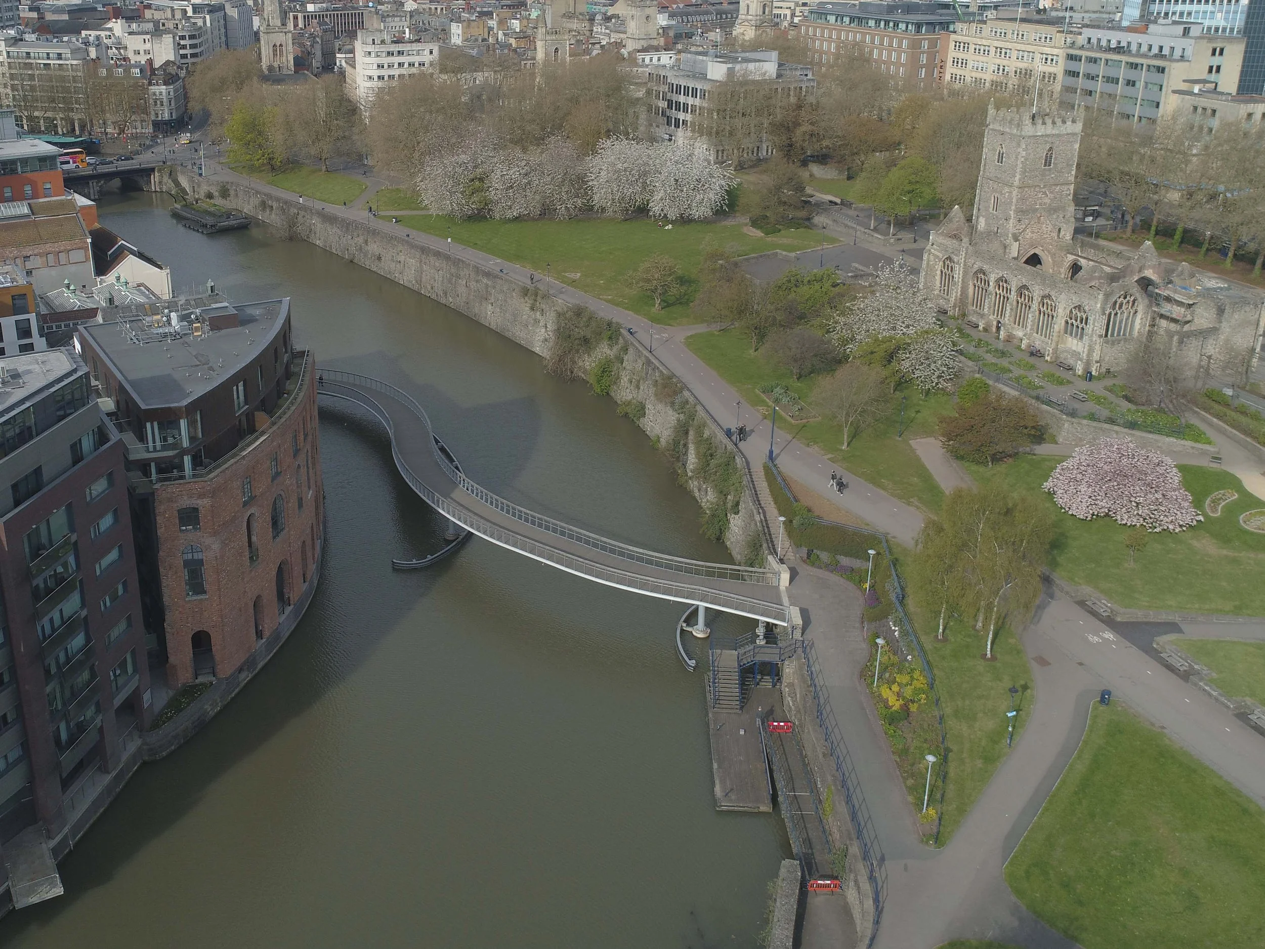 Aerial view of a city park with a river running through it, featuring a modern pedestrian bridge, historic church, blooming cherry trees, and surrounding buildings.