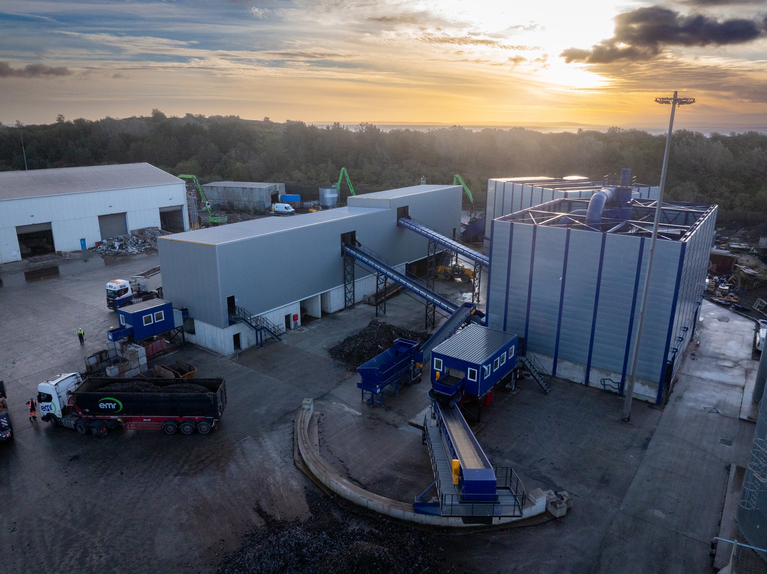 Aerial view of an industrial recycling facility with large buildings, conveyor belts, and trucks, surrounded by trees at sunset.