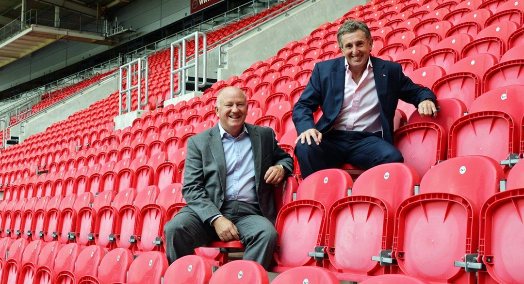 Two men in suits sitting in rows of red stadium seats, smiling.