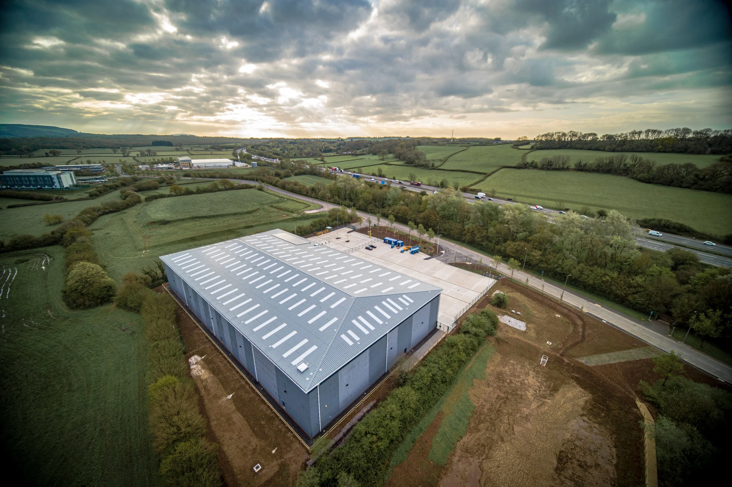 A large industrial warehouse with a metal roof is situated in a rural area surrounded by green fields, trees, and a road with cars, under a cloudy sky.