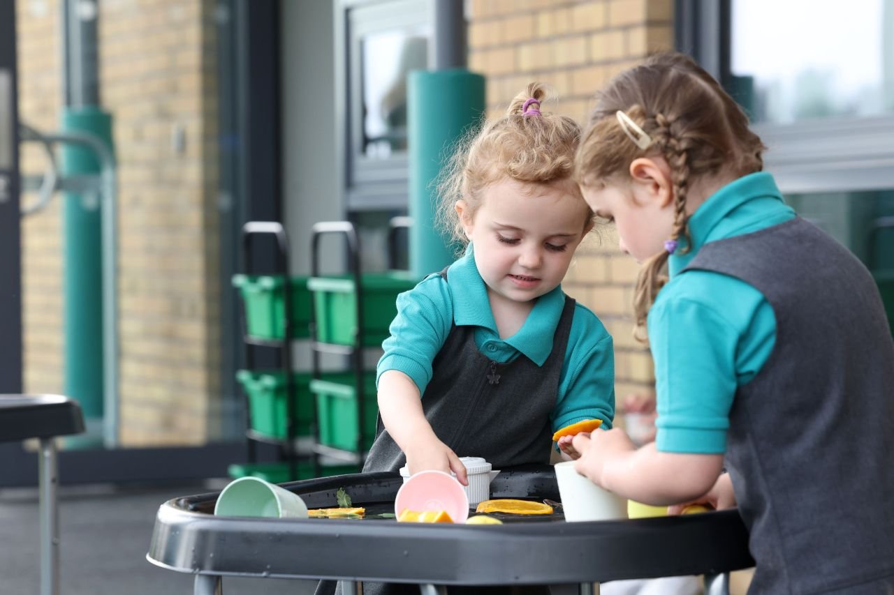 Two young children playing with plastic cups and water at a table outdoors, wearing school uniforms.