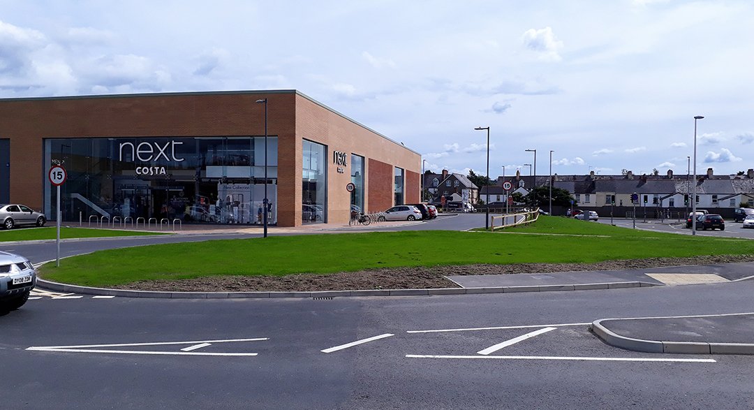 Modern shopping center building with large glass windows and a parking lot in front, surrounded by grass and street lamps, with a few cars parked around.