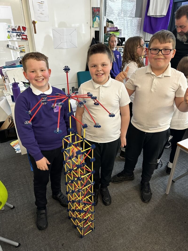 Three boys standing in a classroom with a large structure made of colorful sticks and joints. The structure resembles a skyscraper with various angles and sections. Other children and a classroom setting are visible in the background.