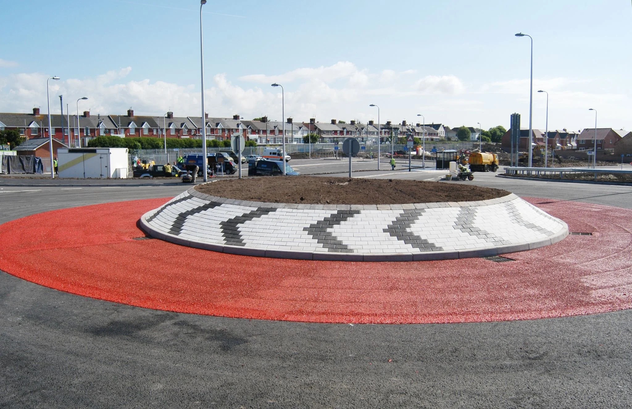 A new roundabout under construction with a decorative black and white arrow pattern on the center island, surrounded by a red textured surface and a paved road.