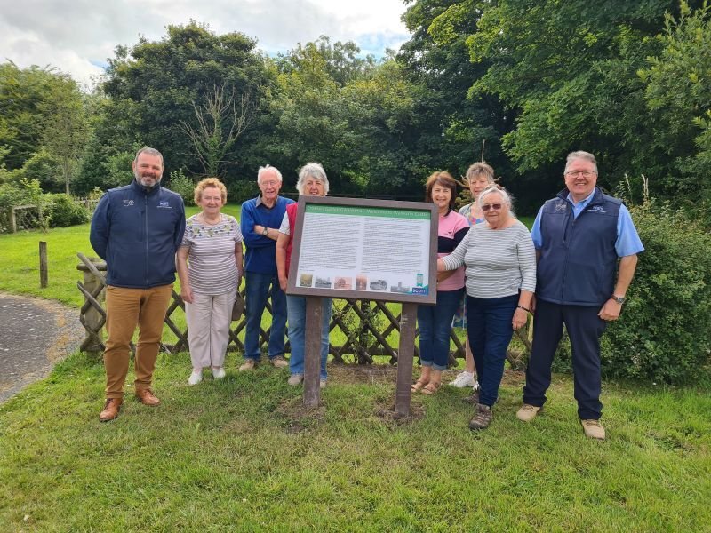 Group of nine people standing outdoors next to an informational sign, with trees and greenery in the background.