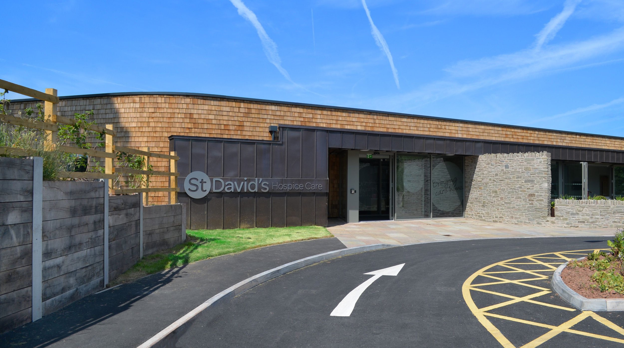 Modern building entrance with "St David's Hospice Care" sign, featuring wood and stone exterior, clear blue sky, and asphalt driveway with white arrow.