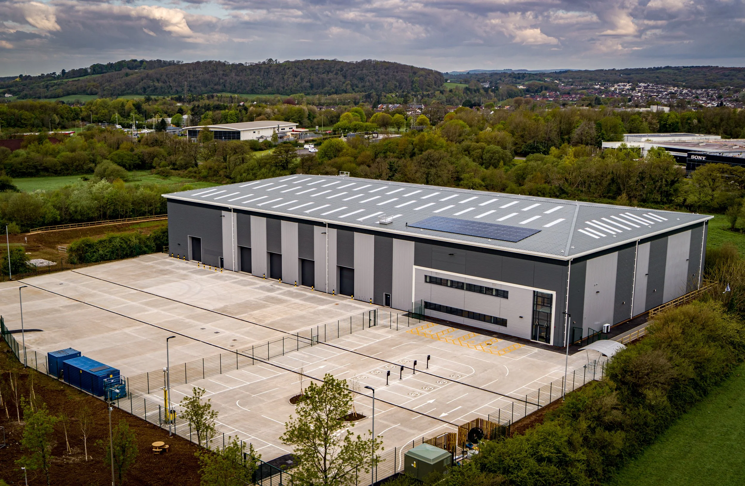 Large industrial warehouse building with multiple roll-up doors, parking lot, and solar panels on the roof, surrounded by greenery and distant hills.
