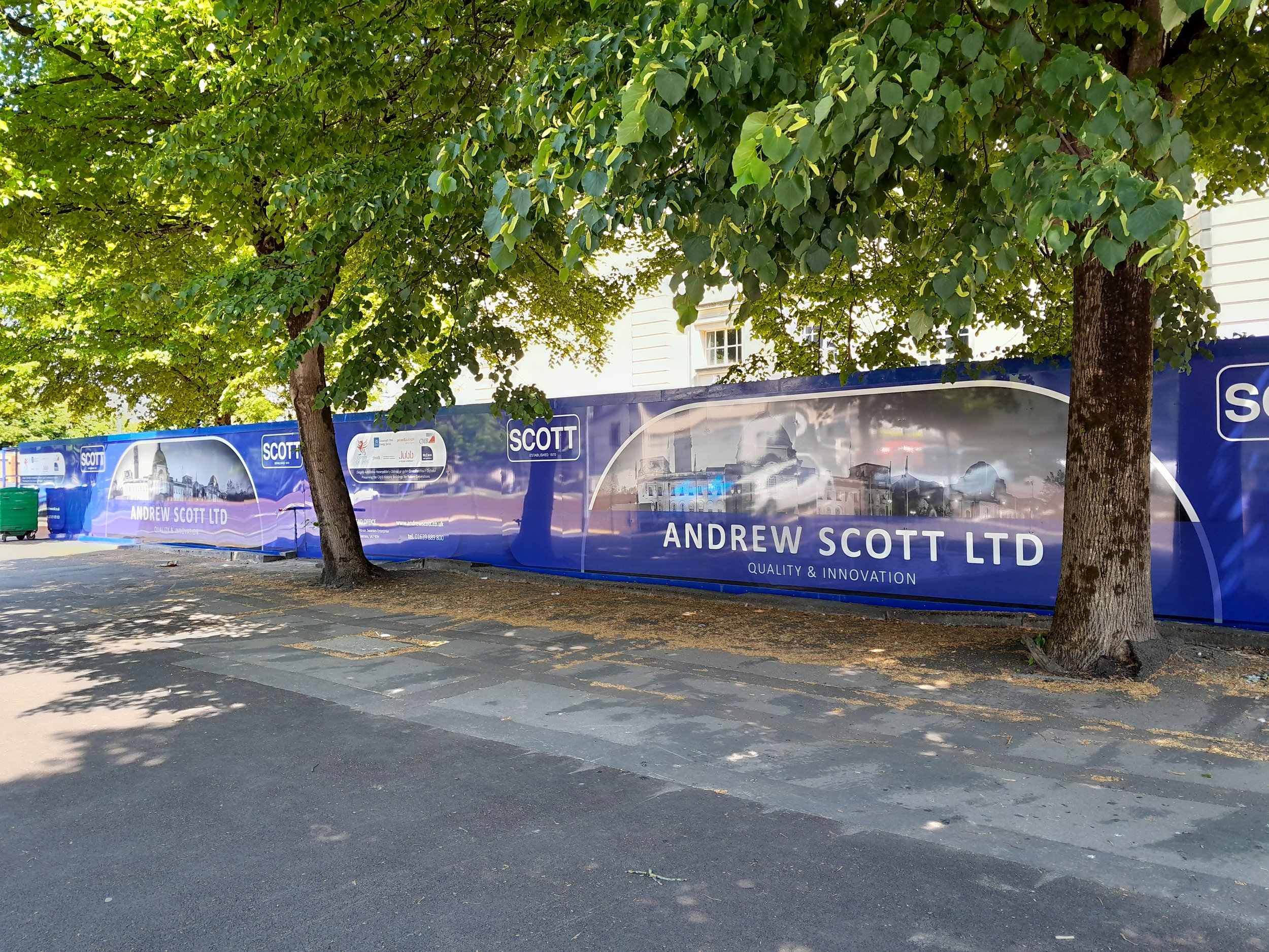 A sidewalk lined with trees and a blue advertising banner for Andrew Scott Ltd, featuring cityscape images and the company logo.