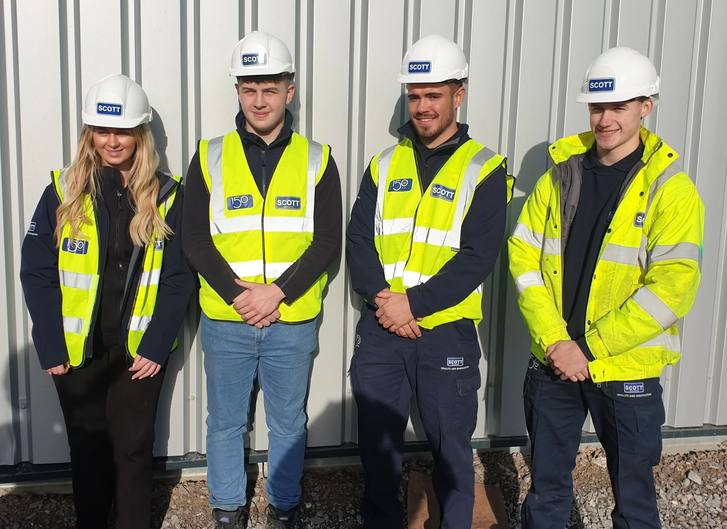 Four people in safety vests and hard hats standing in front of a metal wall, smiling.