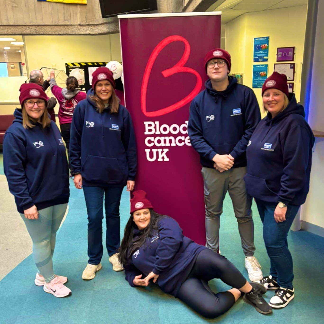 Group of five people wearing blue hoodies and maroon beanies standing in front of a pink banner that reads 'Blood Cancer UK'. One person is lying on the floor. They are in an indoor space, possibly a lobby or reception area.