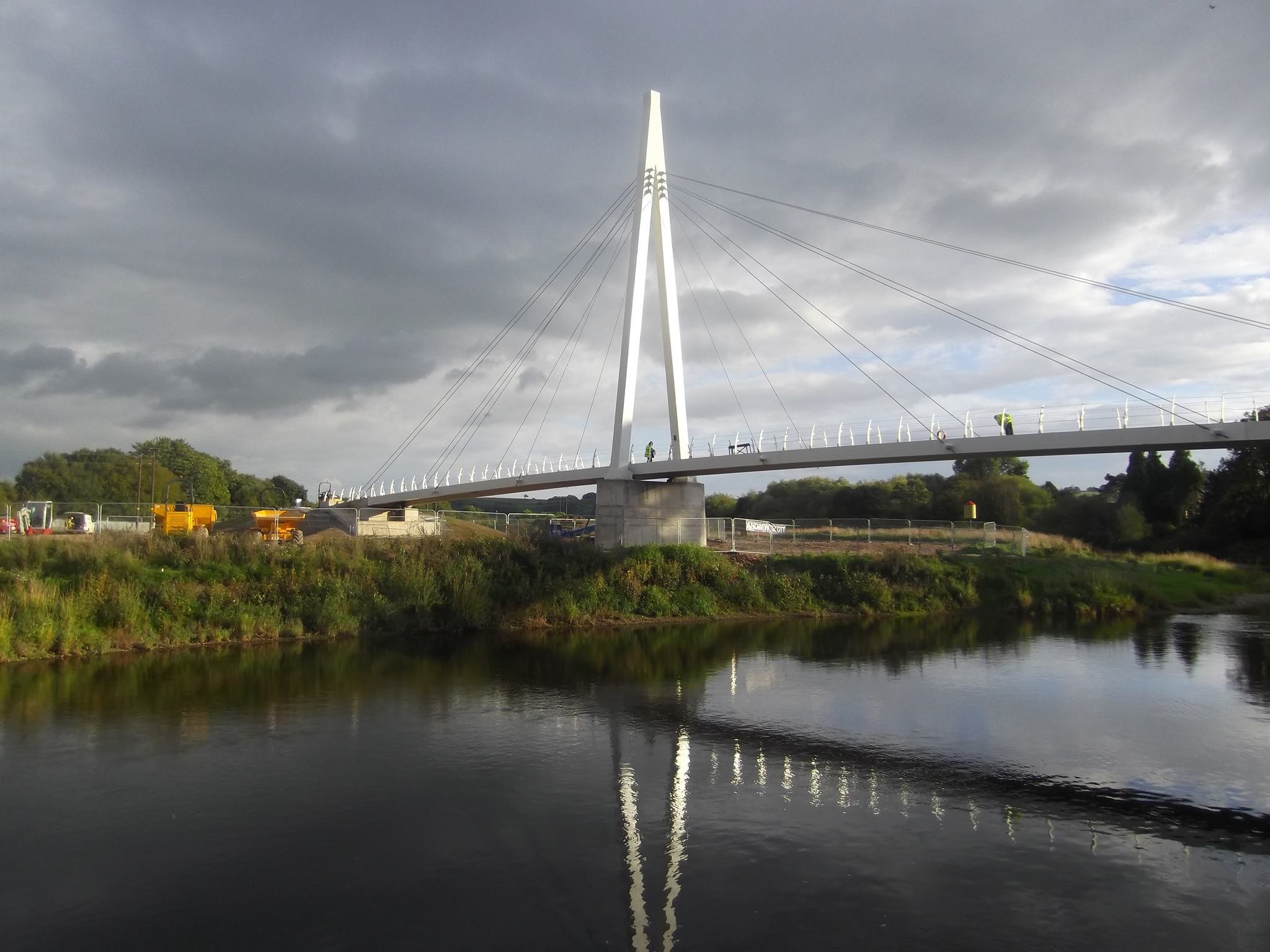 Modern white cable-stayed bridge with a tall pylon over a river, with a cloudy sky in the background and construction vehicles visible on the far side.