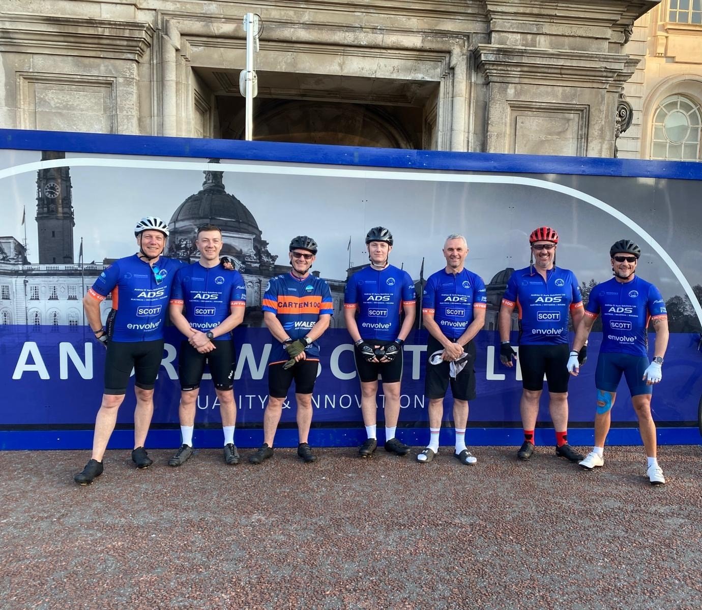 Group of seven men dressed in blue cycling jerseys standing side by side in front of a large sign with the cityscape in the background.