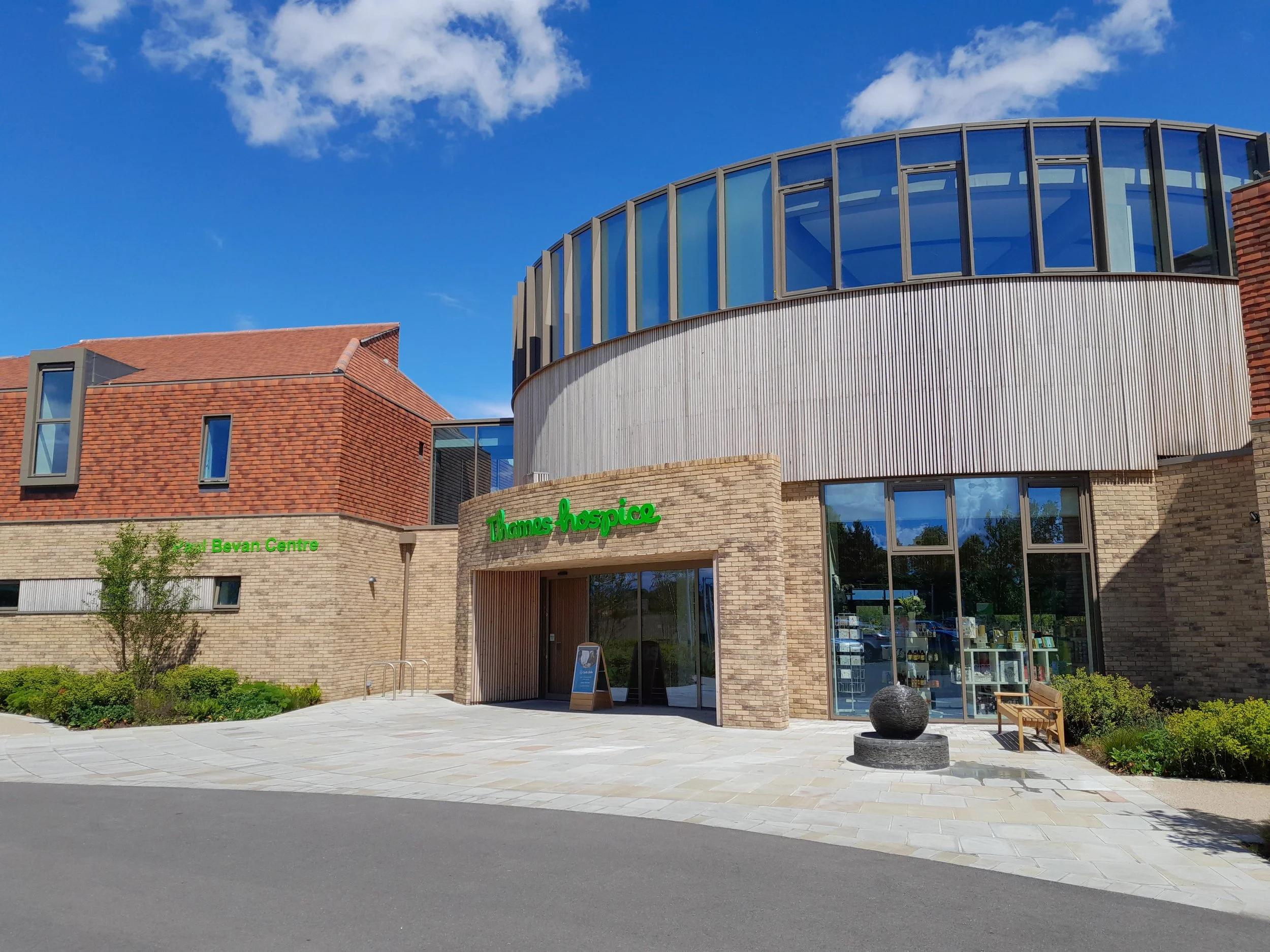 Modern brick and glass building with "Thomas Hospice" and "Bevan Centre" signage, blue sky, and a circular sculpture outside.