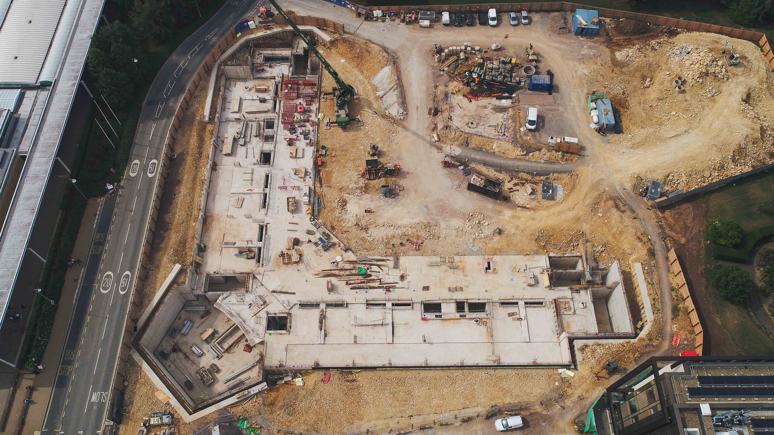 An aerial view of a construction site next to a curved road, with buildings under construction, construction equipment, and piles of dirt and materials.