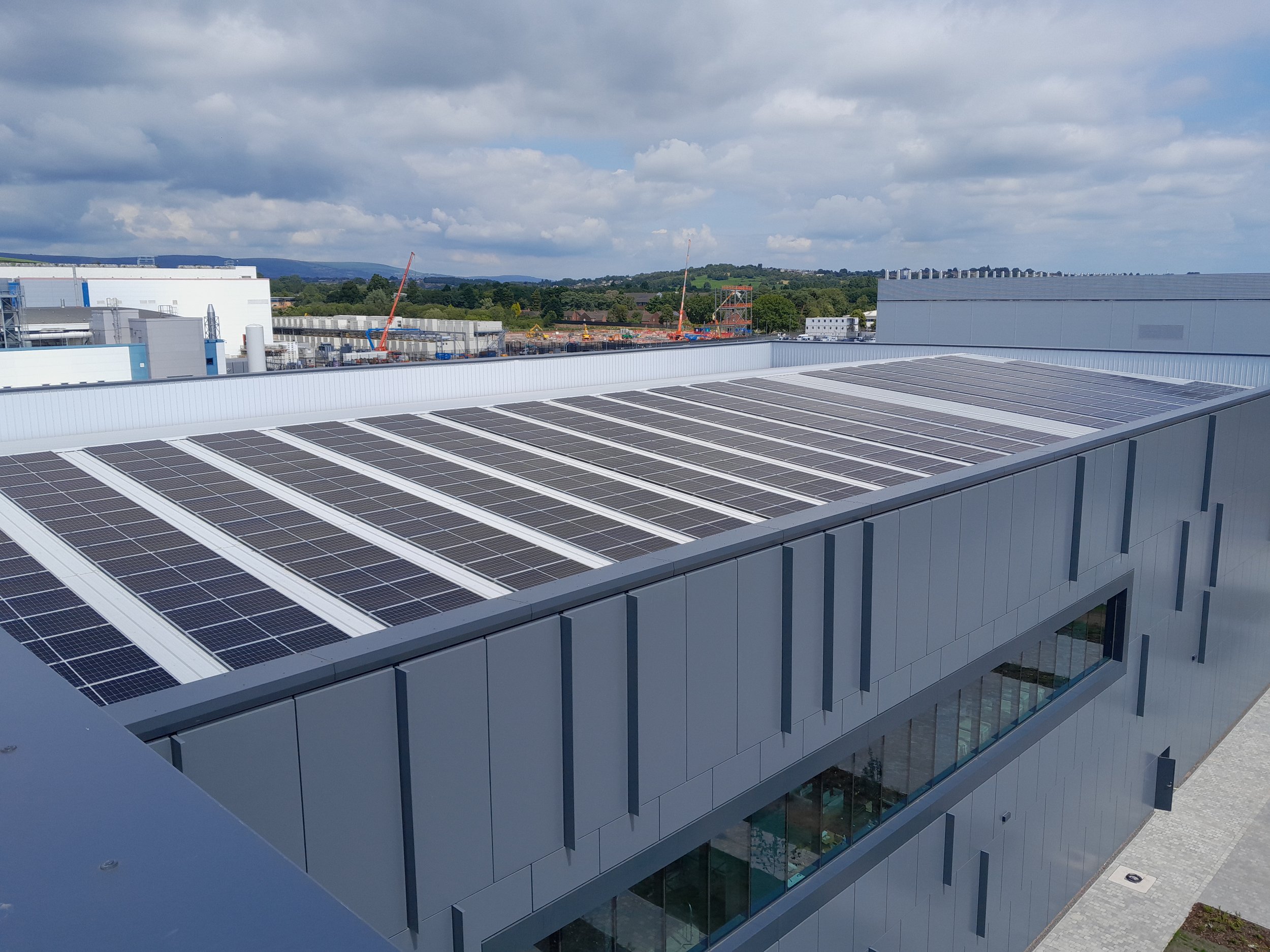 A modern building with solar panels on the roof, located in an industrial area with other buildings and a cloudy sky in the background.