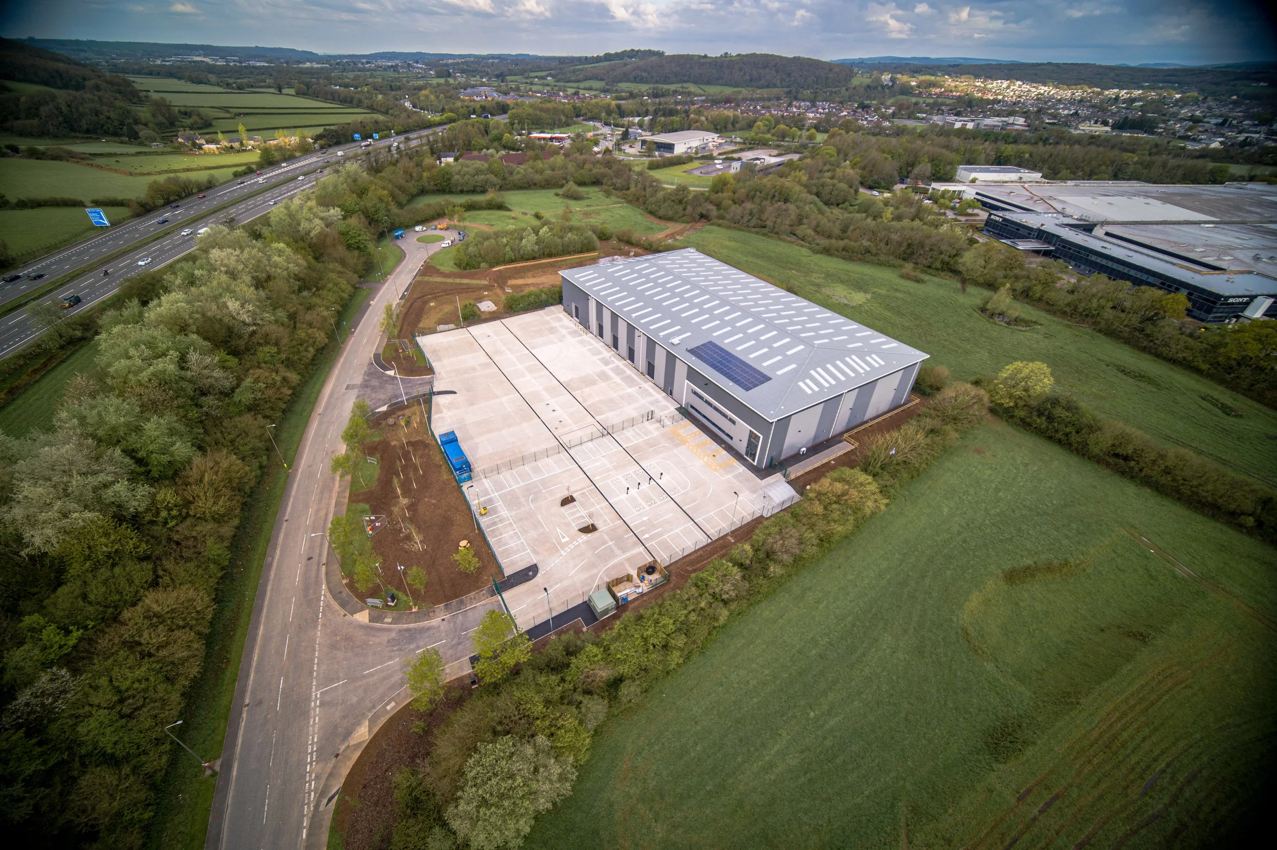 Aerial view of a modern warehouse building with solar panels on the roof, surrounded by a parking lot, green fields, and nearby roads.