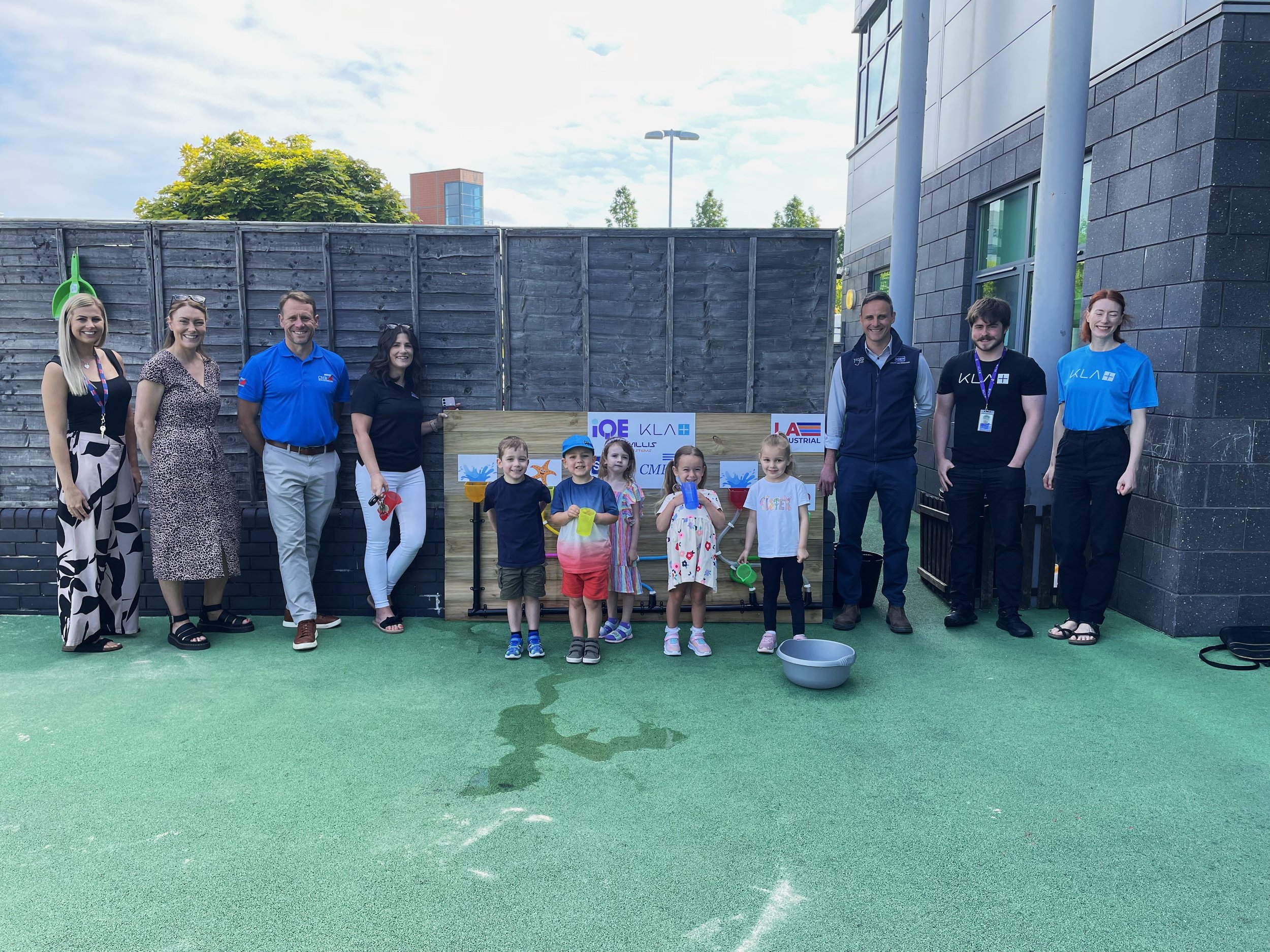 Group of children and adults standing outside on a green surface, some holding water toys, with a wooden and metal fence backdrop, a building to the right, and a cloudy sky.
