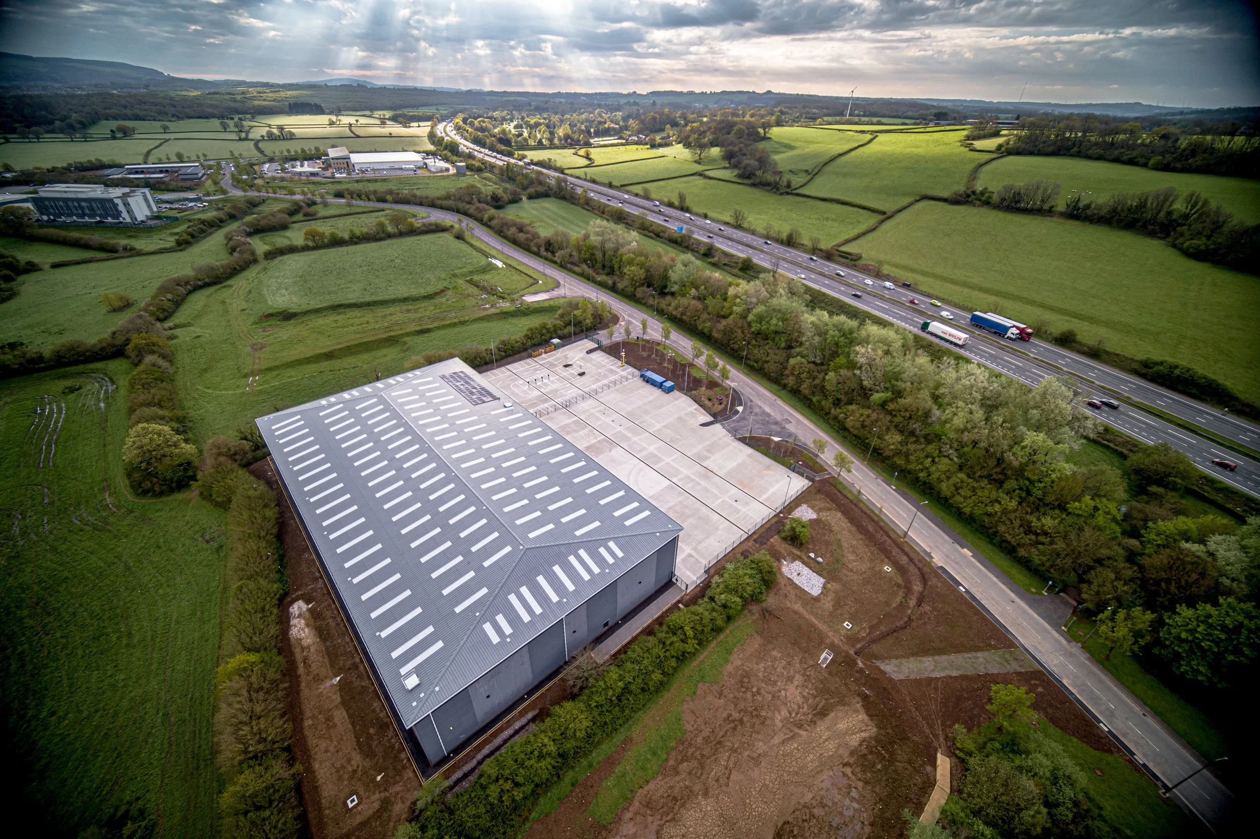 An aerial view of a large industrial building with a metal roof, surrounded by green fields, a parking lot, and a highway with multiple lanes and trucks, under a cloudy sky.