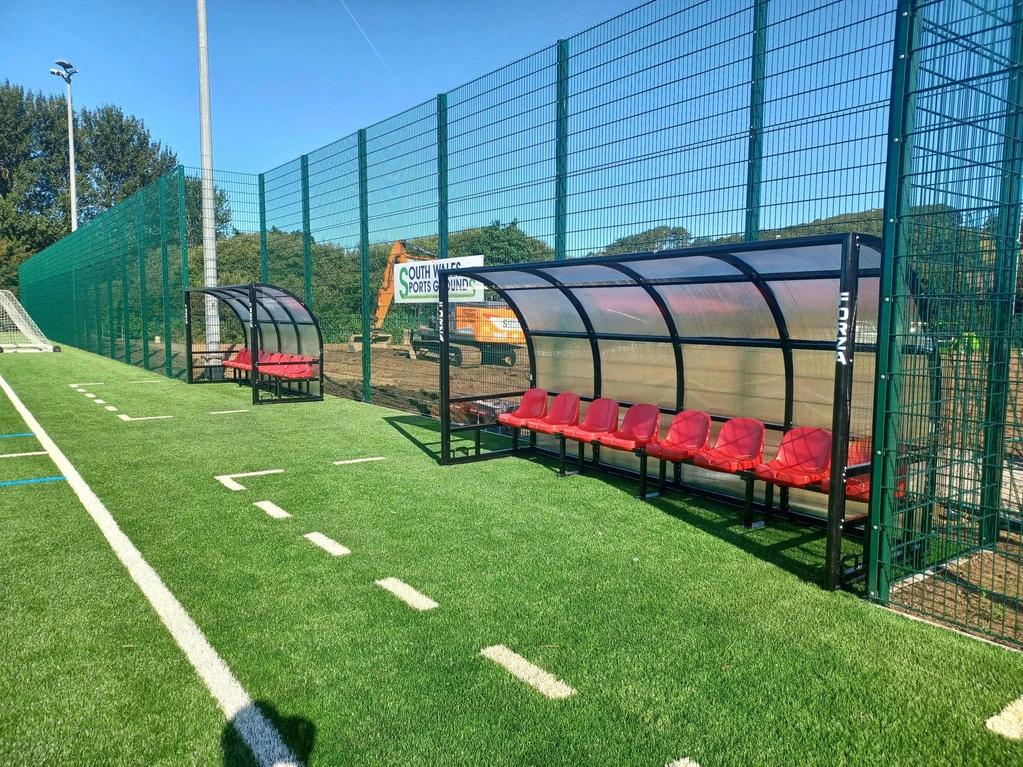 Soccer field with a sideline bench area, red chairs inside a shelter, fenced sports ground under a clear blue sky.