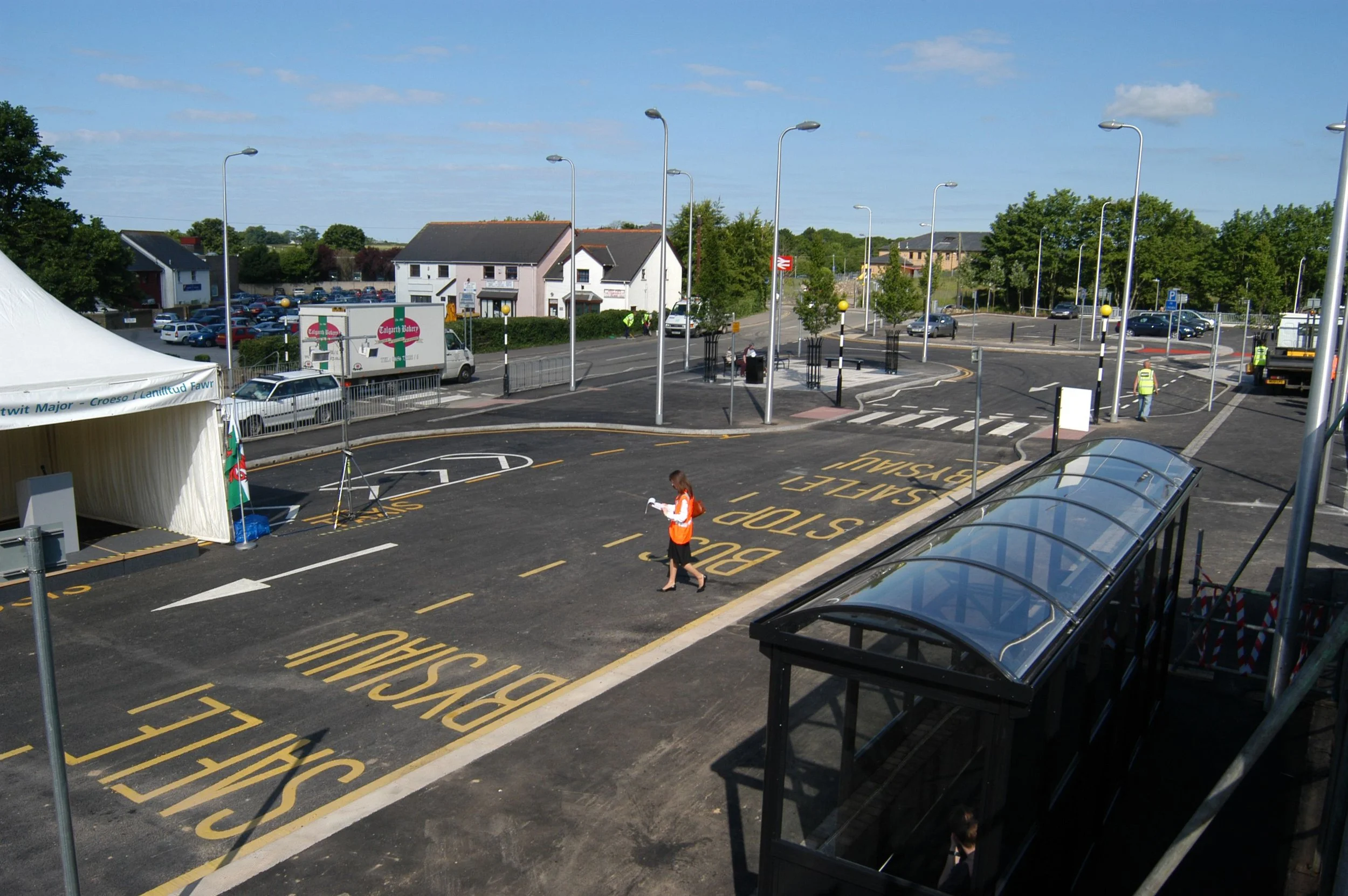 View of a parking lot under clear sky with several marked bus stops, a tent, people walking, and buildings in the background.