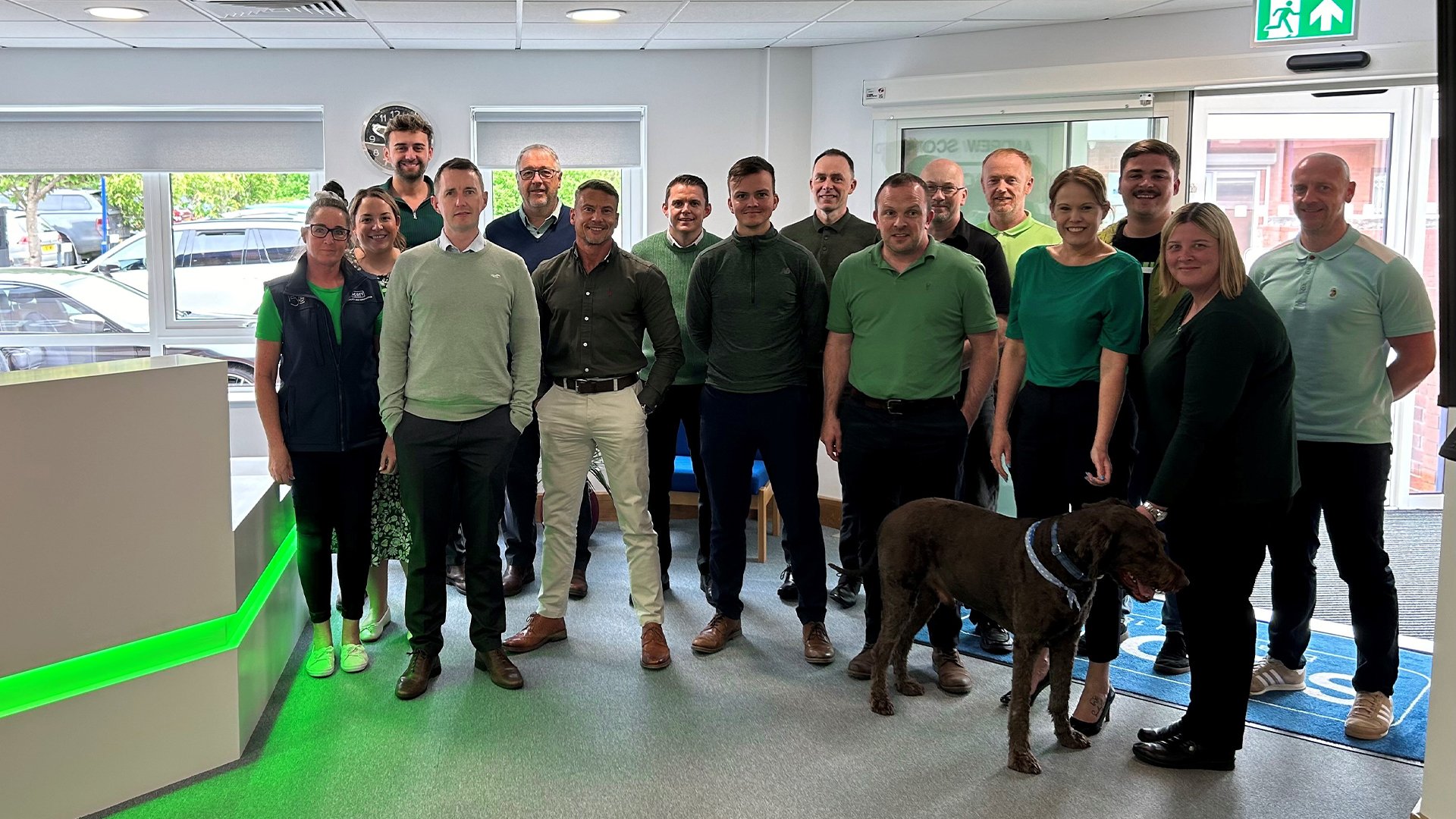 Group photo of people in a room, all wearing green clothes, standing near a reception desk with green lighting, alongside a large brown dog. The room has white walls, large windows, and a clock on the wall.