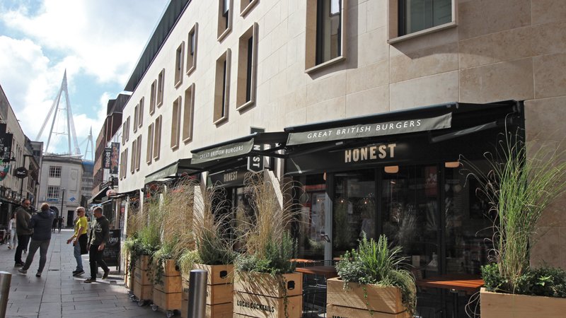 Street scene with a restaurant named Honest displaying "Great British Burgers" on the awning. The outdoor seating is adorned with potted plants. Pedestrians walk along the cobblestone path, and a modern building with a triangular structure is visible