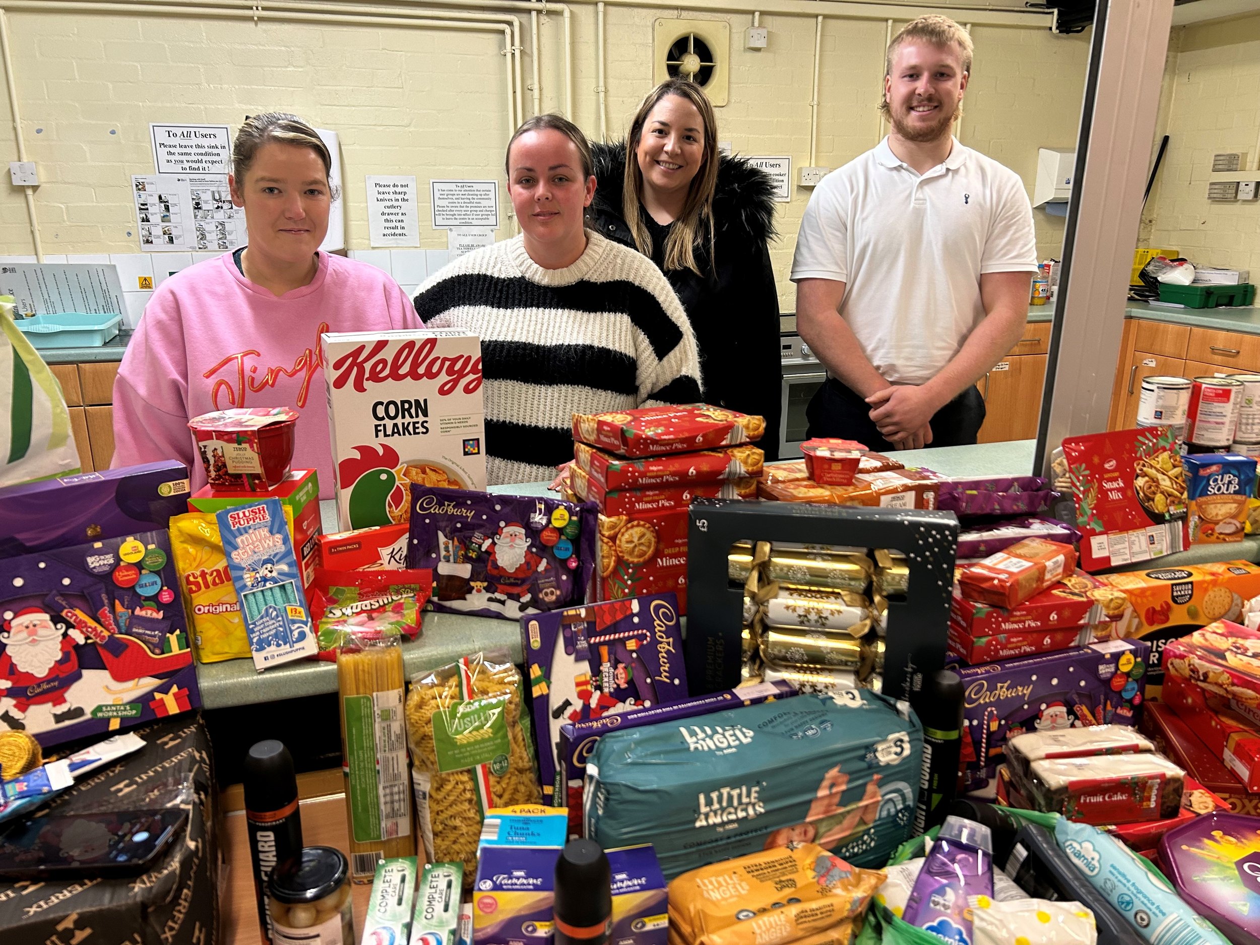 Four young adults standing behind a table filled with various snacks and holiday treats in a kitchen.