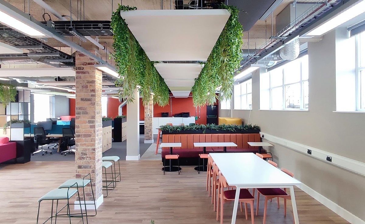 Modern open-plan office interior with hanging greenery, wooden flooring, white tables, colorful chairs, and exposed brick columns.