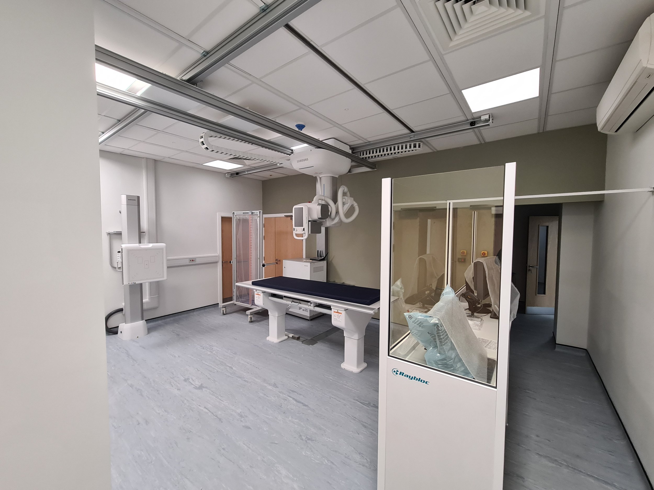 Empty modern X-ray room with medical equipment, including an X-ray machine, table, and protective barrier.
