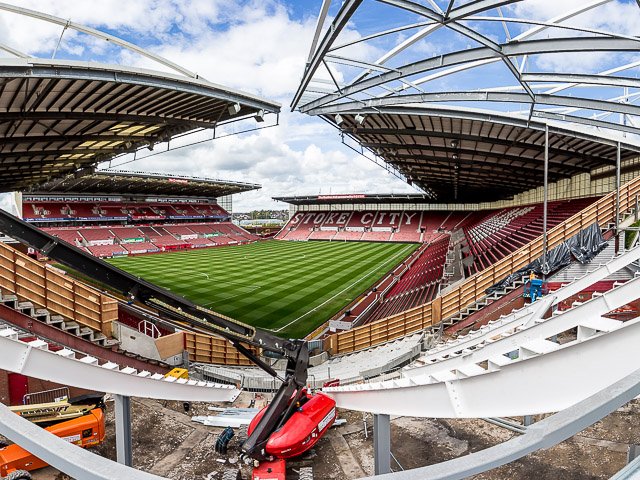 Interior of a soccer stadium under construction or renovation with a view of the field, seating, and partially completed roof structure.