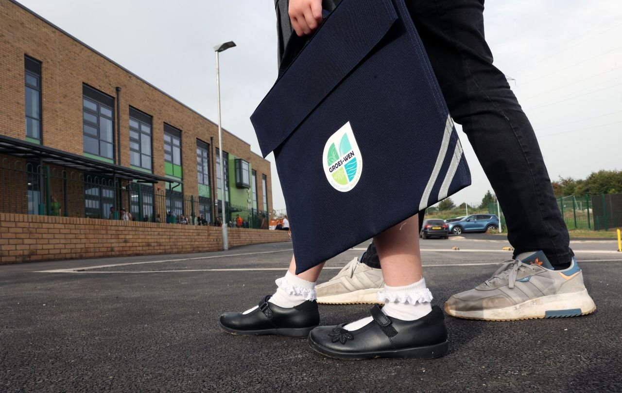 Person holding a blue bag with a school emblem, walking with another person in sneakers, in front of a brick school building.