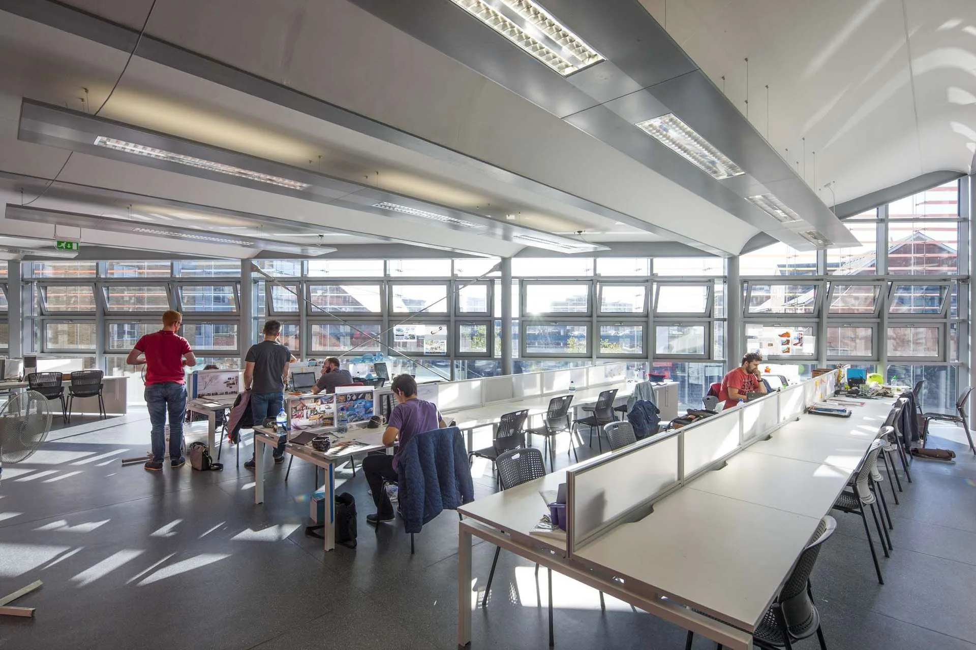 Open office space with several employees working at desks near large windows, some standing and some seated, with sunlight streaming in.