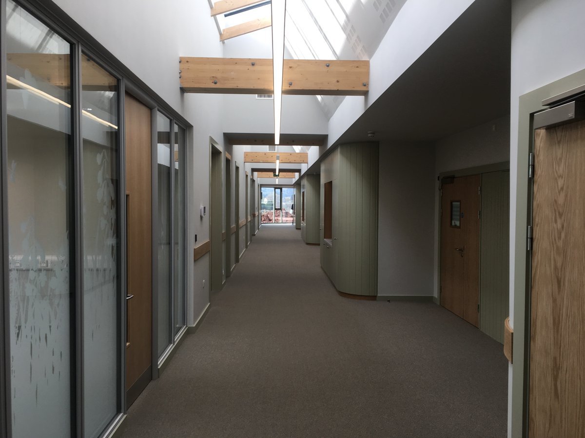 An indoor hallway with glass doors on the left, green and beige walls, wooden beams on the ceiling, and large windows at the far end showing an outside view.