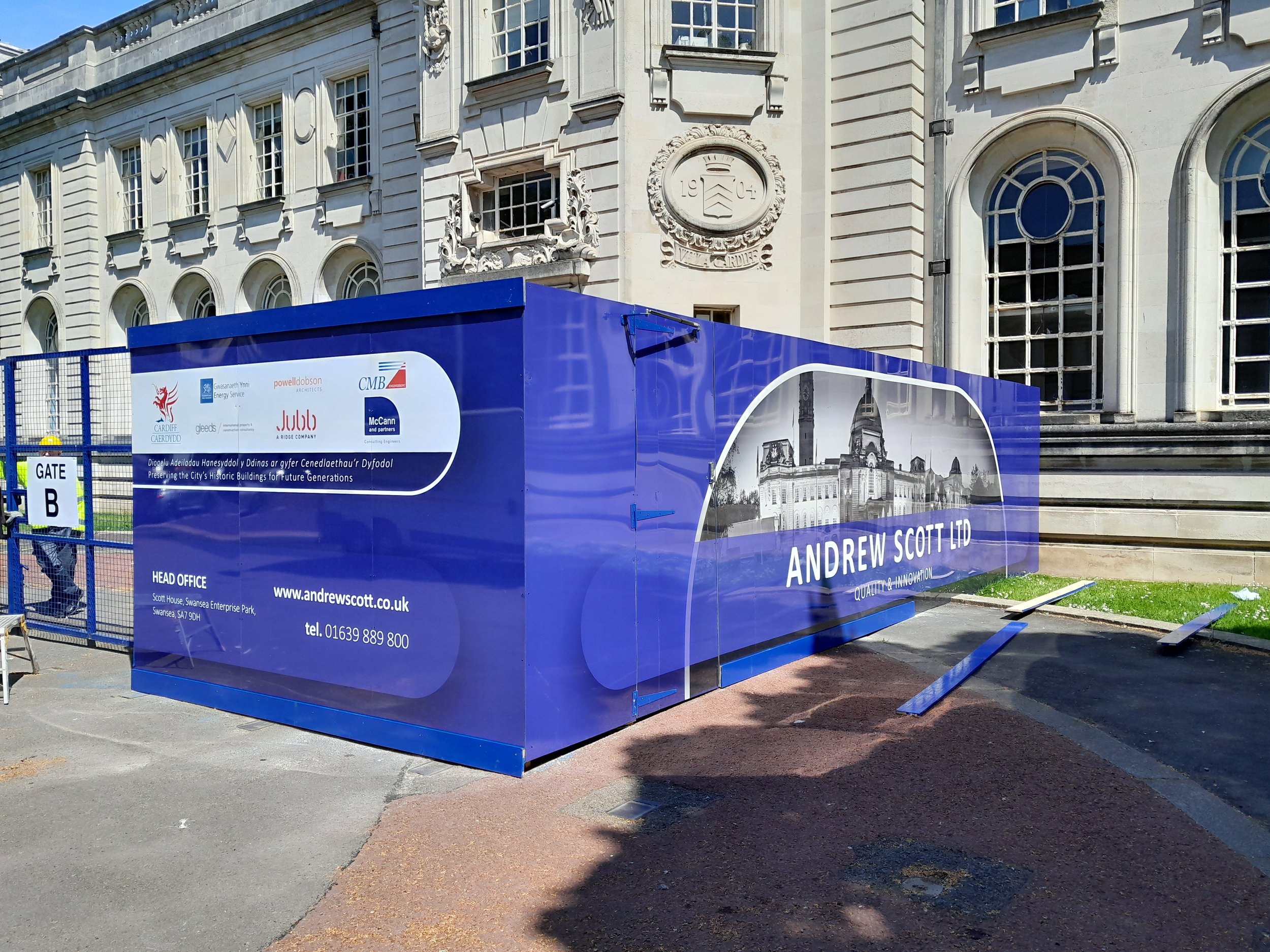 Blue construction site enclosure with branding and a black-and-white image of a historic building, located in front of a large classical building with ornate details.