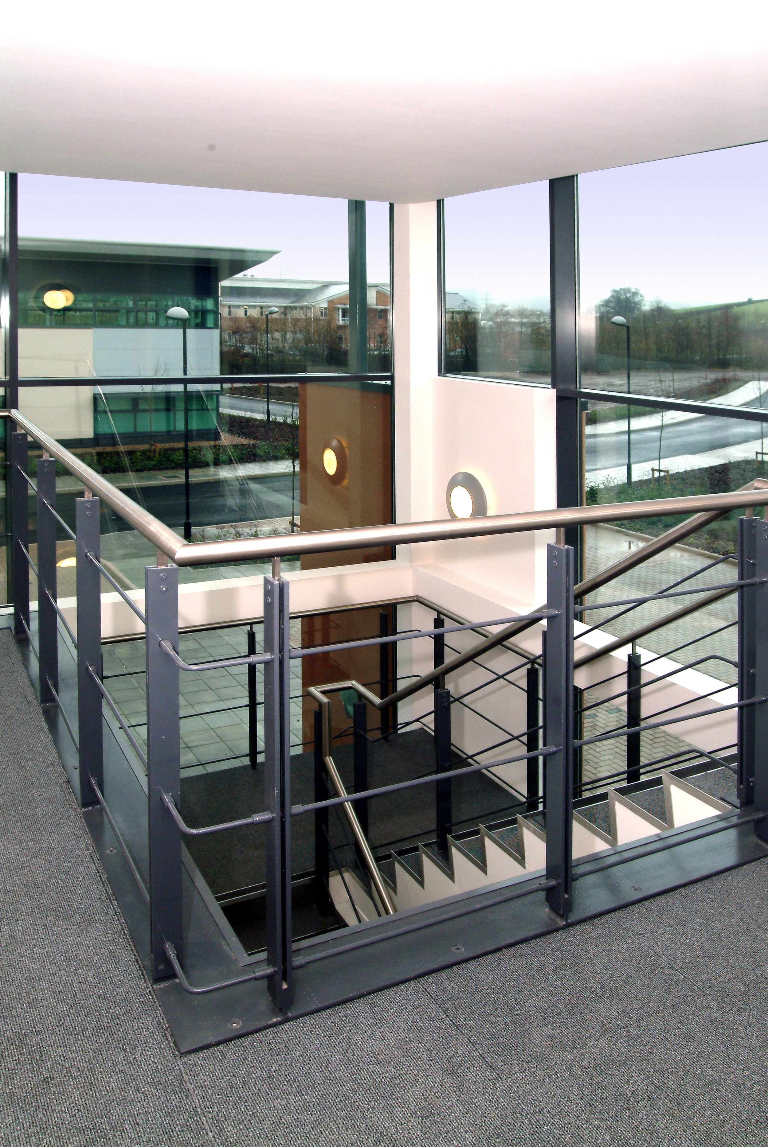 Interior of a modern building with a staircase, metal railings, and large glass windows showing an outdoor view.
