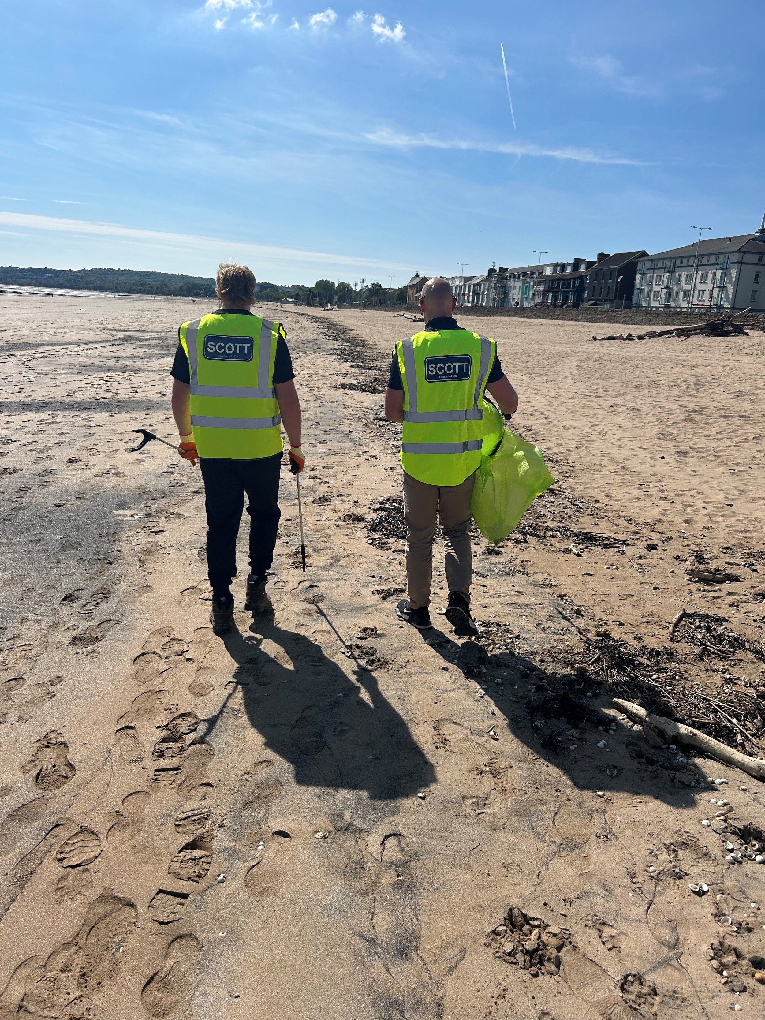 Two people wearing neon yellow vests marked 'SCOTT' are cleaning a sandy beach, walking away from the camera toward the horizon, with footprints and some debris visible on the sand.