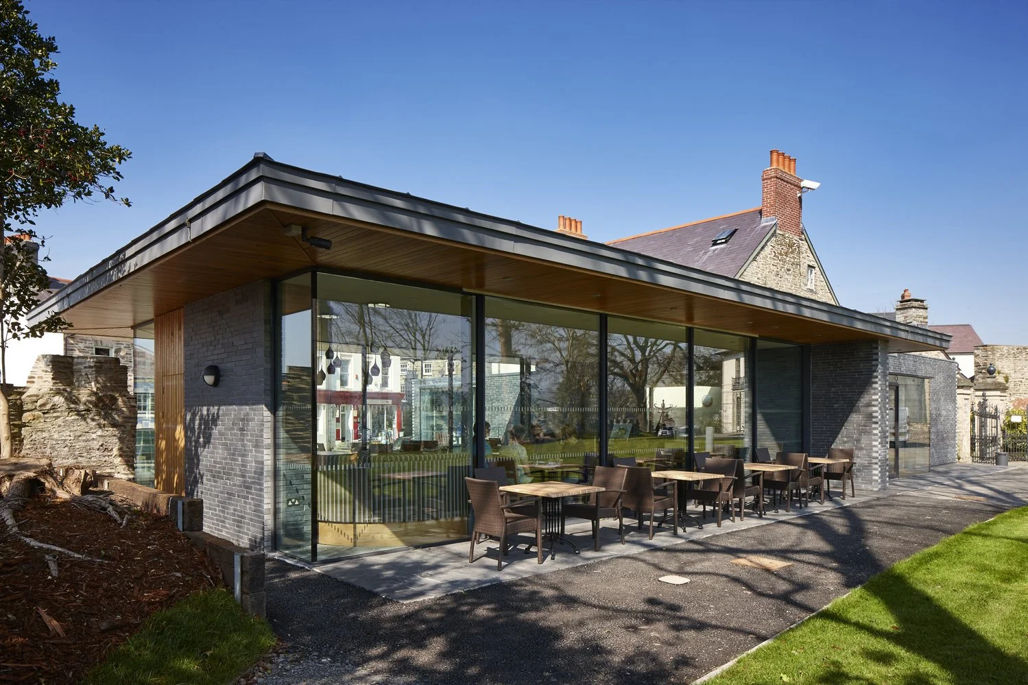 Modern glass-fronted building with outdoor seating, adjacent to old stone structure, under a clear blue sky.