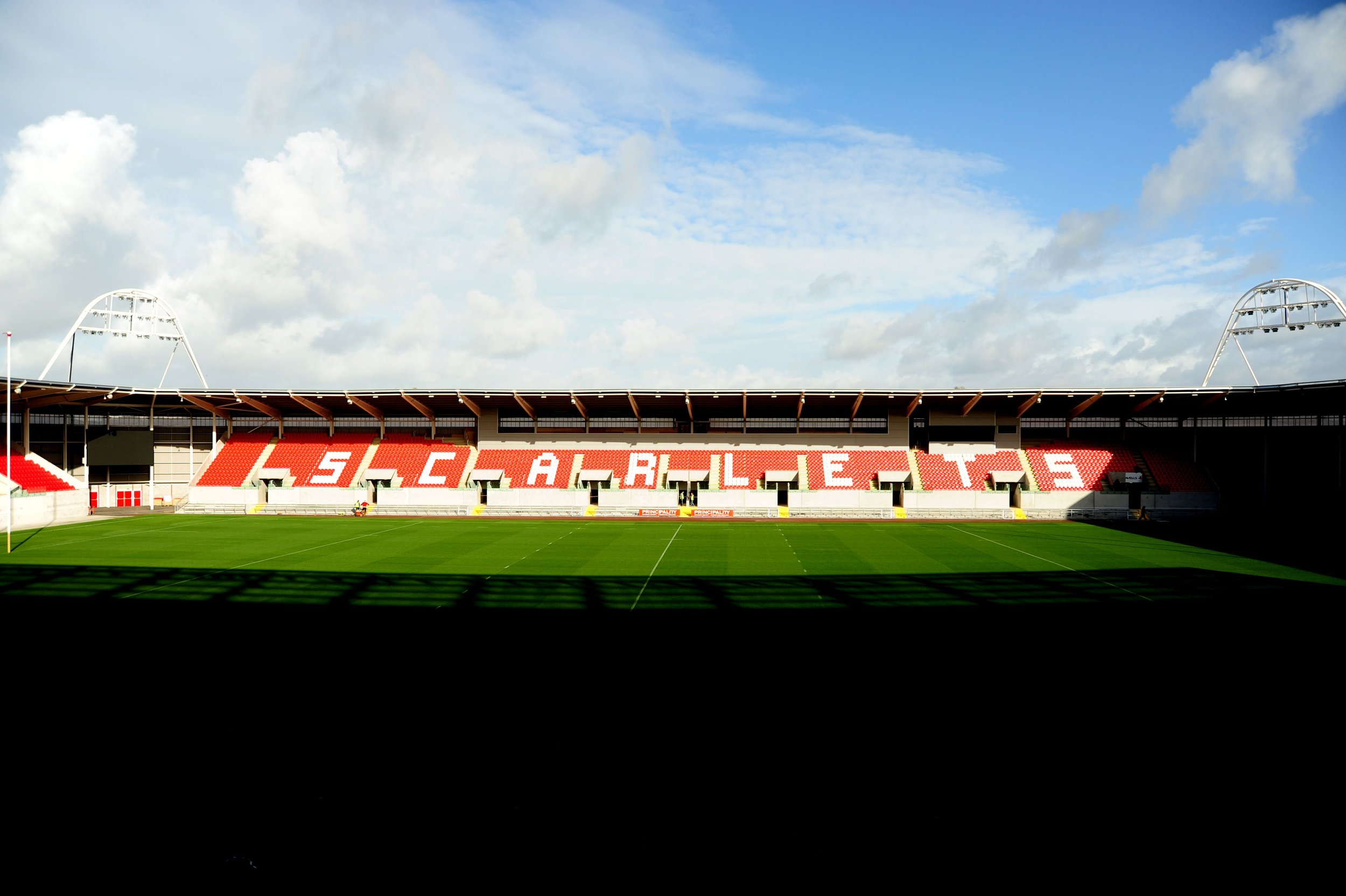 Empty sports stadium with "Scarlets" on red and white seating, green field, and blue sky.
