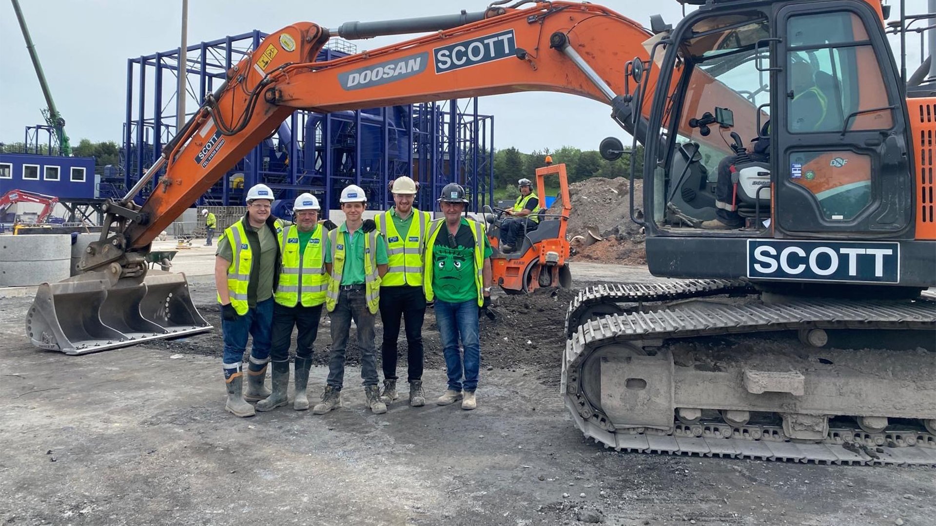 Construction site with four workers in high-visibility vests standing in front of an orange excavator marked 'Scott,' with an operator in the cab, and blue structures in the background.
