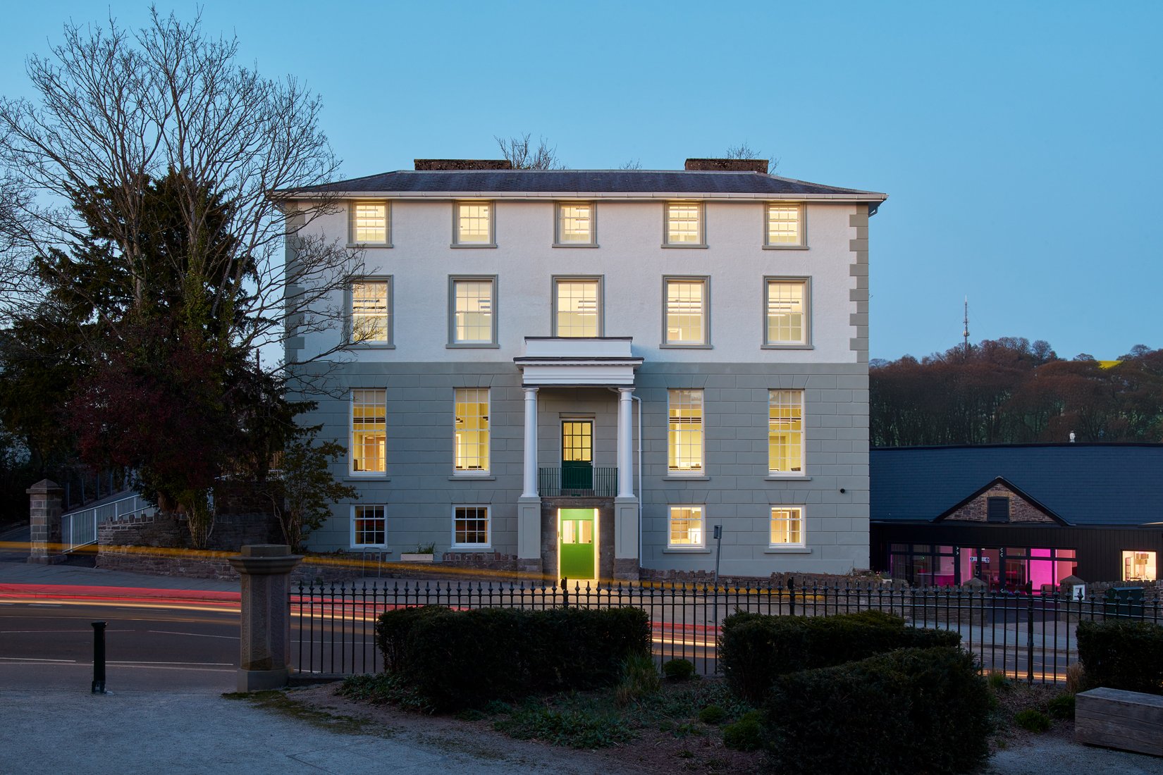 Facade of a historic white and gray building with illuminated windows at dusk, surrounded by trees and a road in front. The building features a small entrance porch with columns.