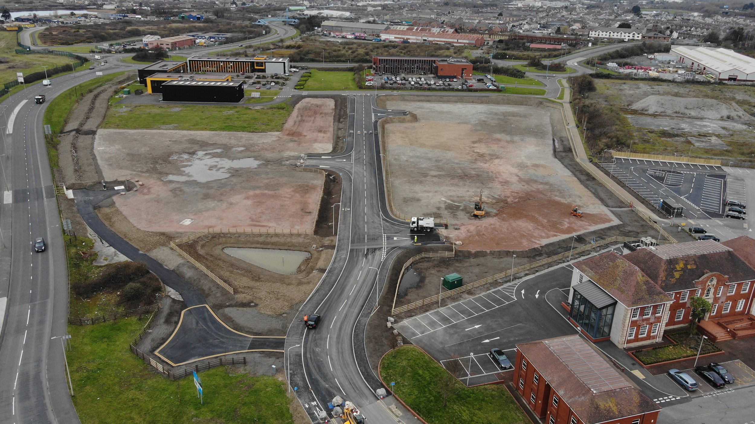 Aerial view of a construction site with newly paved roads, parking lots, and surrounding buildings. Open plots of land, machinery, and grassy areas are visible.
